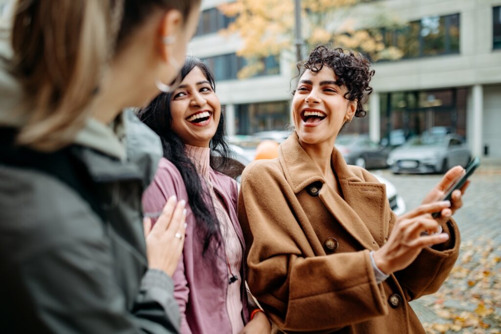 Drei Freundinnen stehen im urbanen Umfeld und schauen gemeinsam auf ein Smartphone